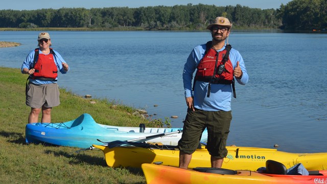 Two people wearing lifejackets stand on a bank alongside several kayaks