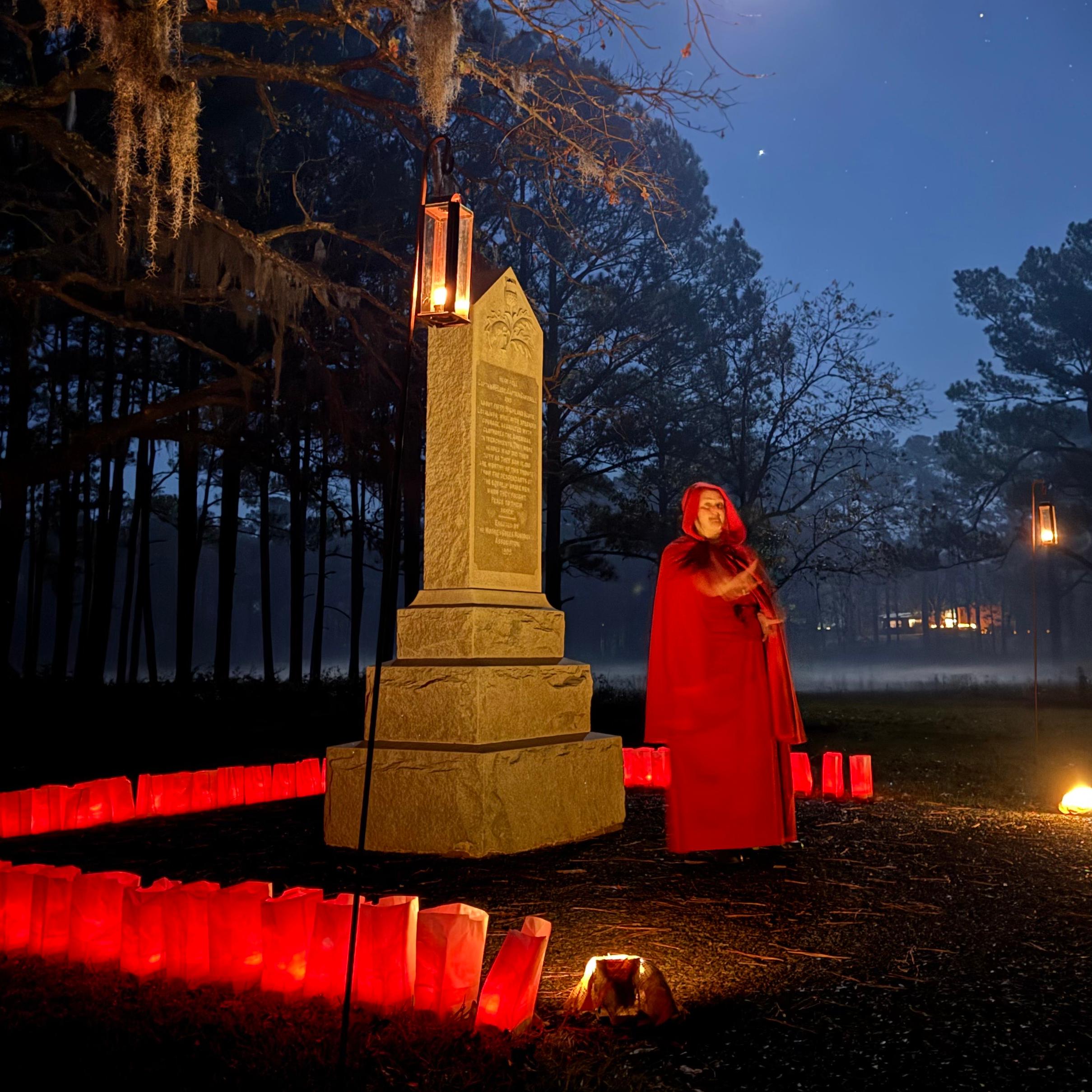 Woman in a red cloak standing in front of a monument surrounded by red luminaries.