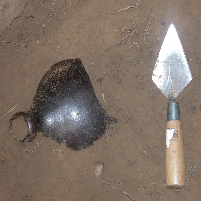 A brown jug is partially exposed in the dirt. It lays next to a trowel.