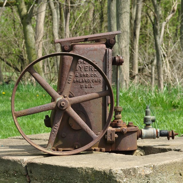 A rusty water pump sitting on a concrete square.