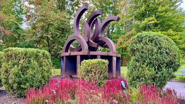 a memorial in rusted bronze with broken olympic rings  with red plants in front