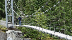 A hiker crosses a long suspension bridge over a rocky river. 