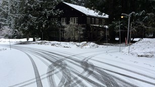Tire tracks cross on a snowy road in front of a wood and stone building.
