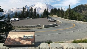 A wayside panel on a low rock wall overlooks a road leading towards a glaciated mountain peak.
