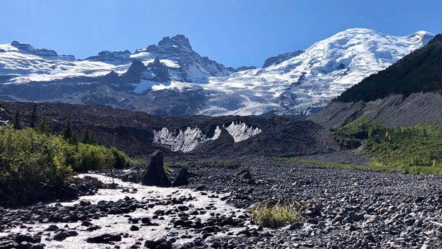 A river flows out of the terminus of a rock-covered glacier on the slopes of a mountain.