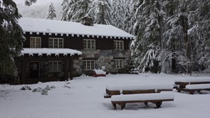 A wood and stone building tucked away in the forest and blanketed in snow. 