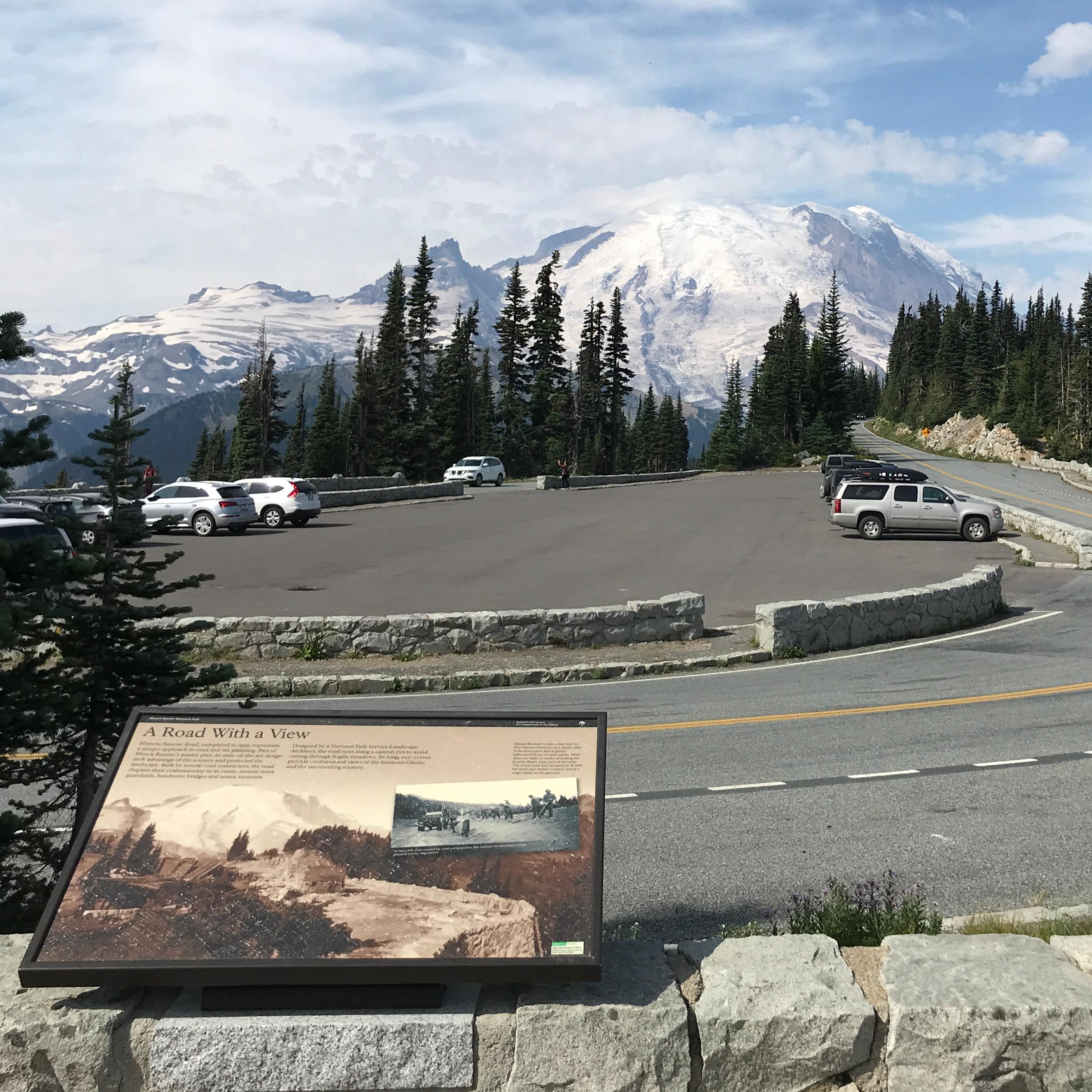 A wayside panel on a low rock wall overlooks a road leading towards a glaciated mountain peak.