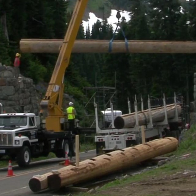 A large truck lifts a large log to span above a road between to rock walls. 