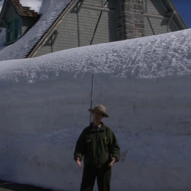 A ranger stands in front of a tall snowbank.