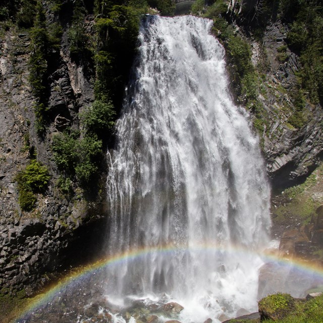 A rainbow arches through the spray of a waterfall cascading down a rocky cliff. 
