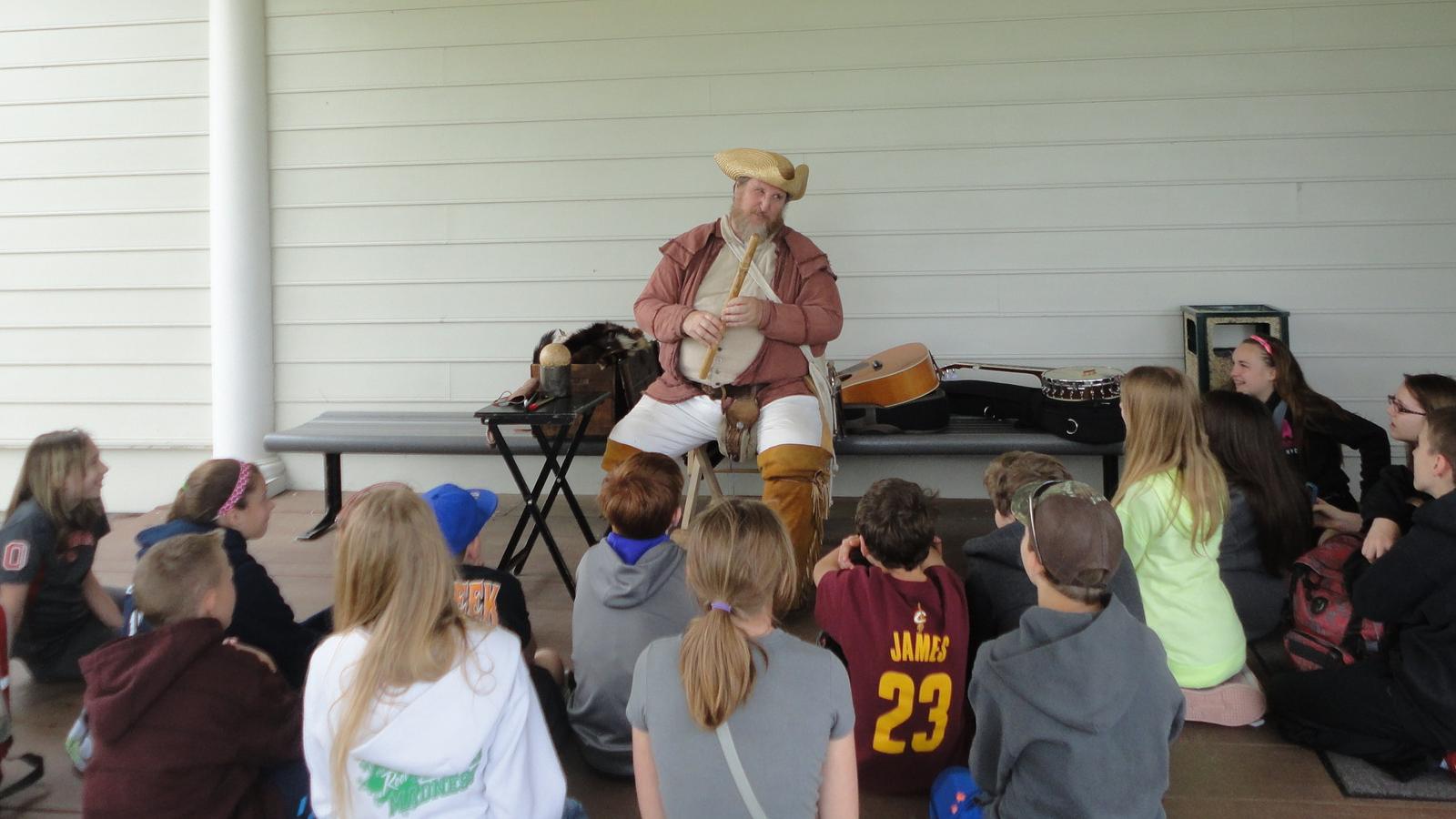 Students sit at the foot of a Man in historic dress hair holding a Fife.