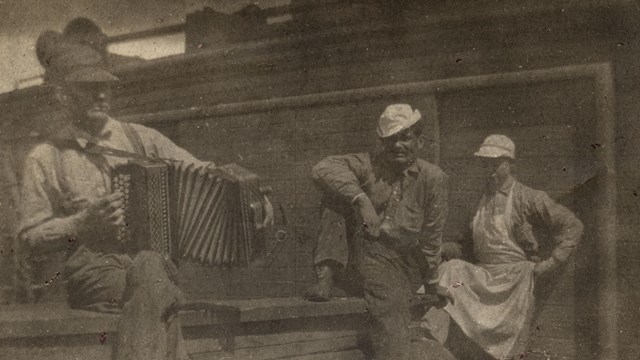 three men sit on a deck of a vessel with one playing the accordian. 