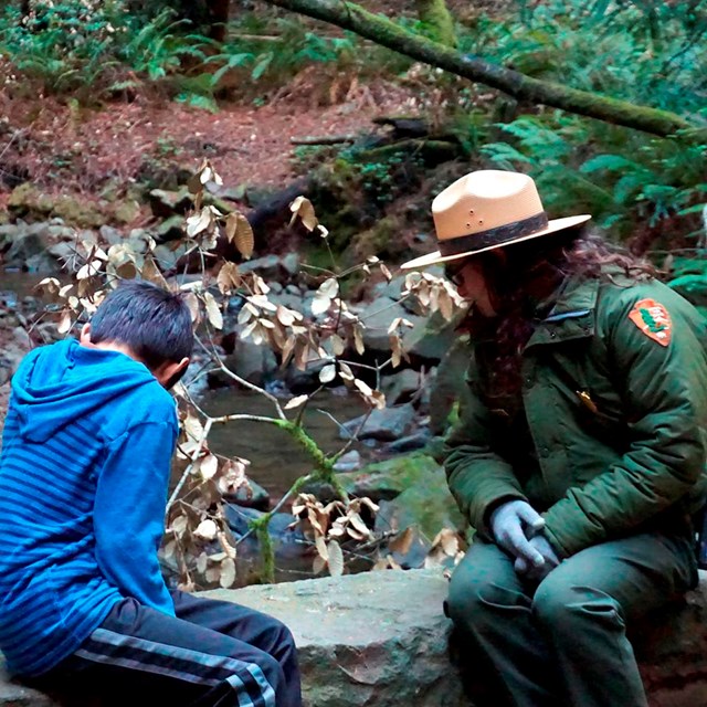 A ranger and a child sit on a bridge and look at the creek