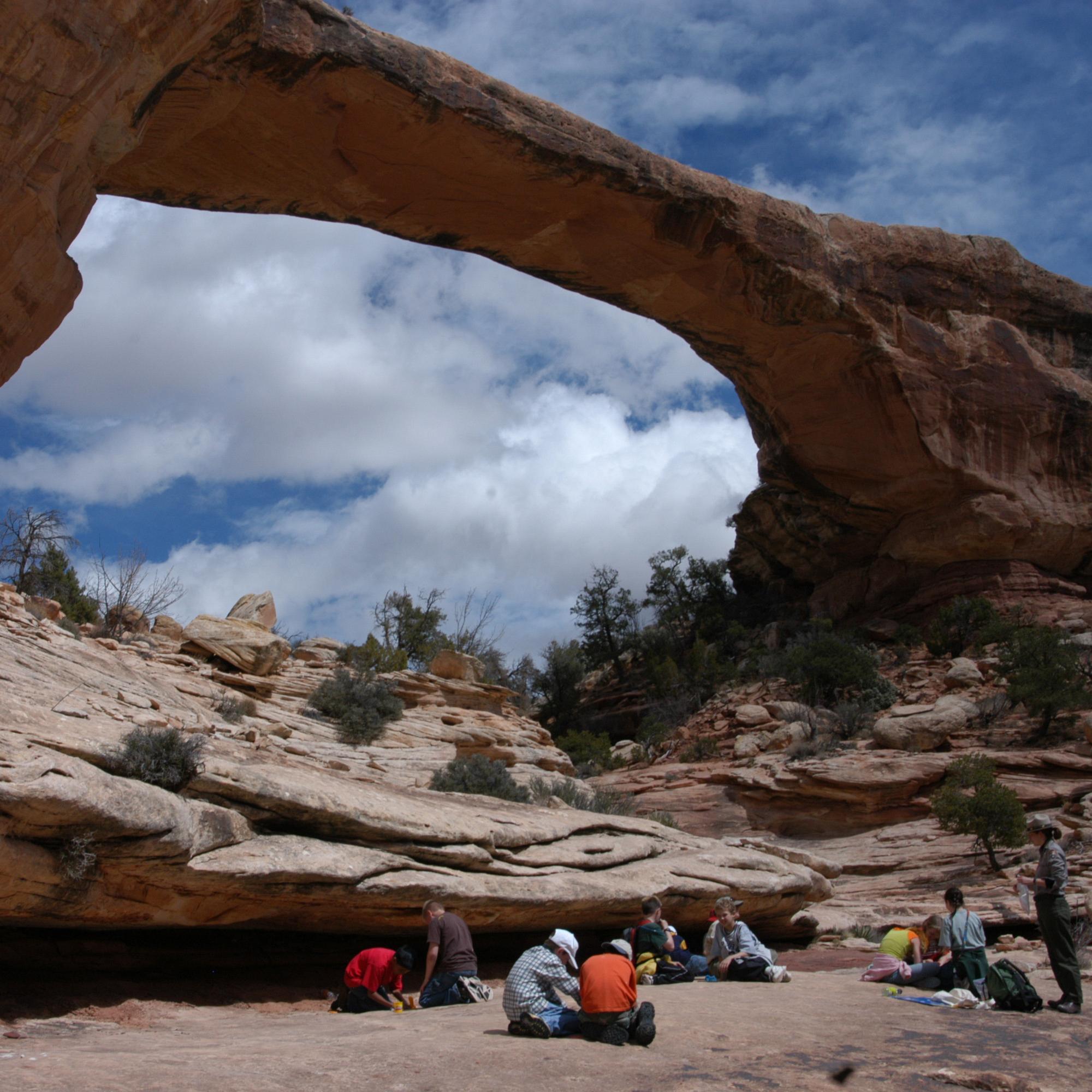 A group of students sit below a large sandstone rock bridge. Blue sky and clouds visible above.