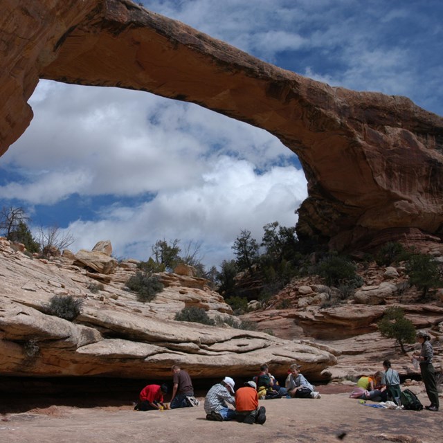A group of students sit below a large sandstone rock bridge. Blue sky and clouds visible above.