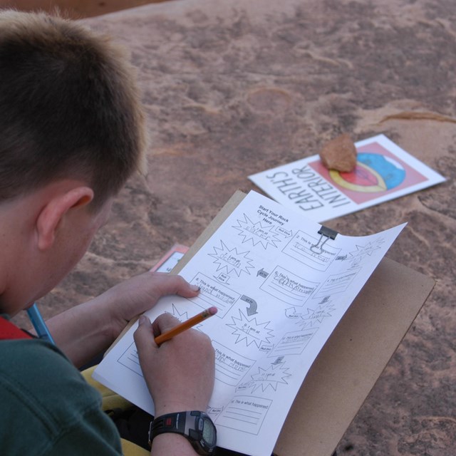 A young person with short hair writes on a clipboard with a worksheet on it. Rocks are behind it.