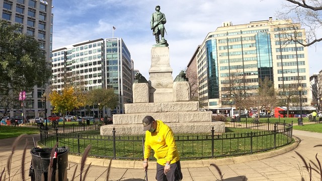 A man in yellow sweeps trash with a statue in the background.&nbsp;