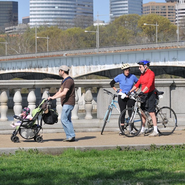 Person walks with child in stroller, two bicyclists stopped on sidewalk, and person walking.