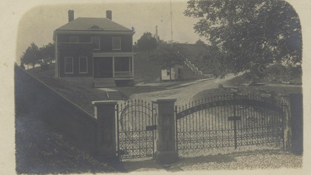 Grayscale photo, entrance to Andrew Johnson National Cemetery with metal gates and two-story house,.