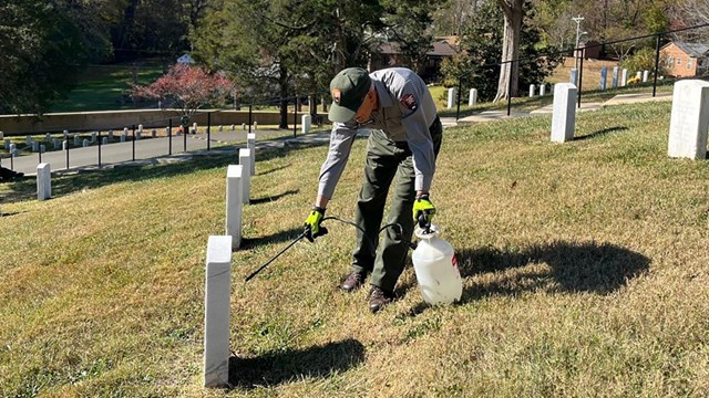 AJ, in NPS uniform, stands in an area of turf and leans to spray the face of a grave marker.