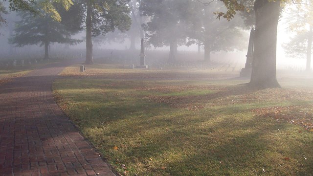 Light filters through fog in a cemetery, surrounding turf, large trees, a brick walk, an headstones