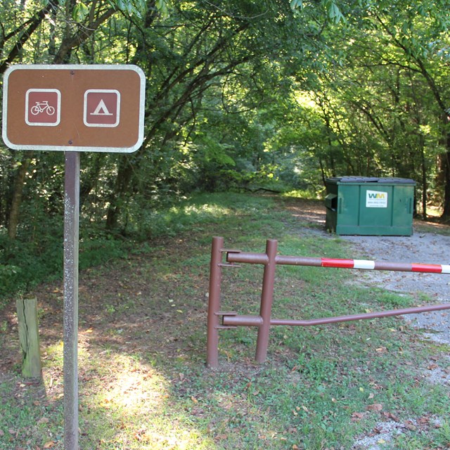 A brown sign stating bicycle camping next to a road blocked to vehicle traffic.