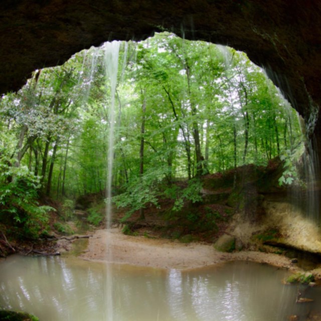 Water drips from a dark natural arch into a muddy stream. 
