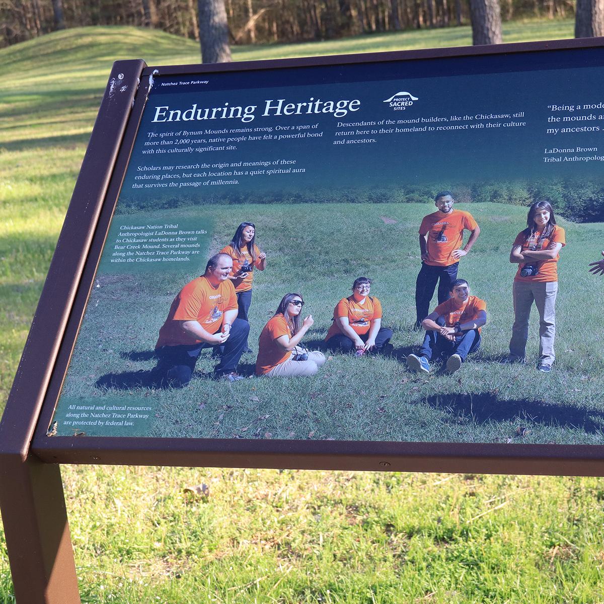 A photo of a sign showing several people in orange t-shirts listening to a speaker.