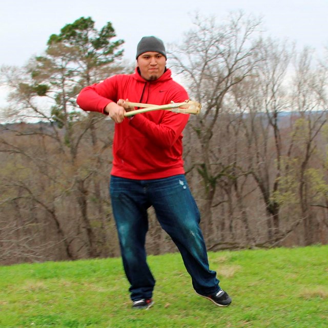A man with tan skin wearing jeans and a red hoodie holds a stickball stick.