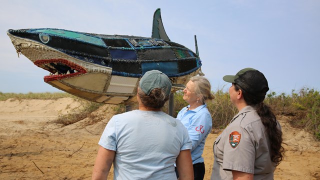 three women stand next to a 14-foot white shark sculpture made out of marine debris