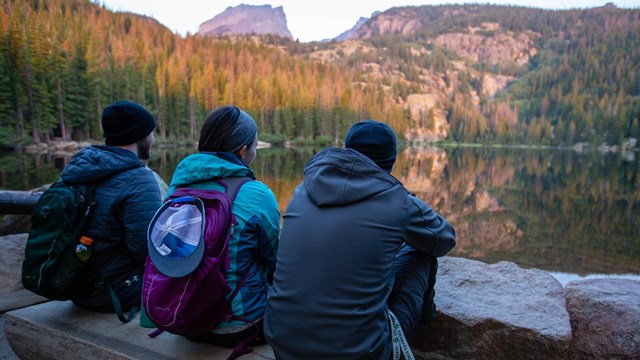 three people dressed in warm clothes sit on a wooden bench overlooking a lake a sunlit trees