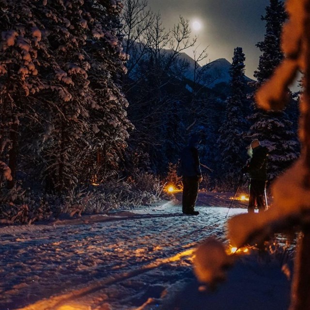 two people on a groomed snowy trail lined with lanterns under a full moon