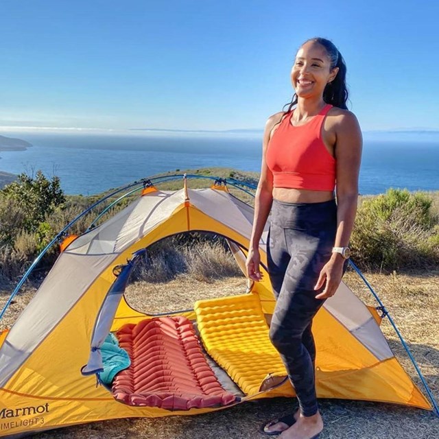 a smiling young woman stands next to a tent on a bluff overlooking the ocean