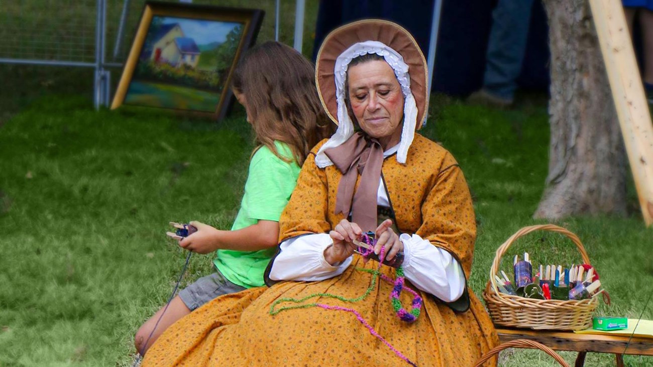 Woman in historic dress crochets beside a child at an outdoor event with baskets of supplies nearby.
