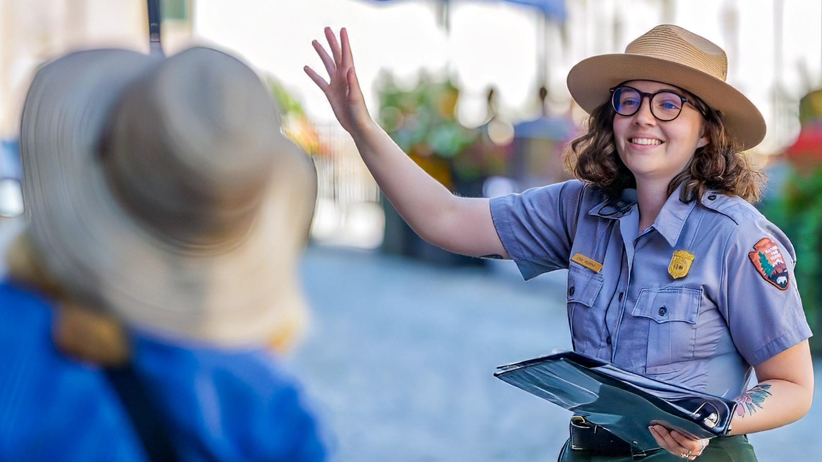 A park ranger smiles and waves while leading a tour outdoors, holding a clipboard and speaking to vi