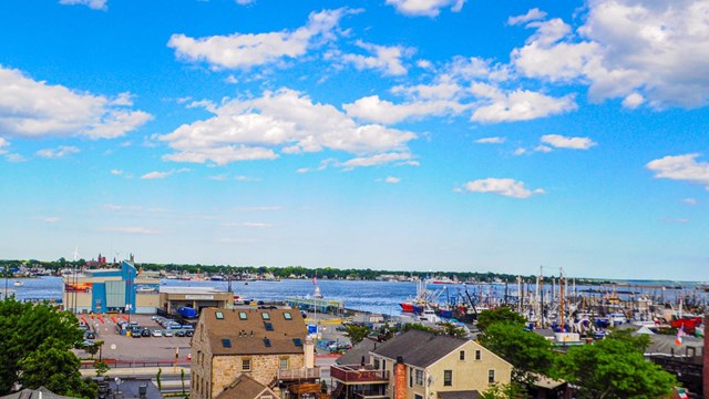 View of New Bedford harbor with fishing boats, historic buildings, and a bright blue sky with clouds