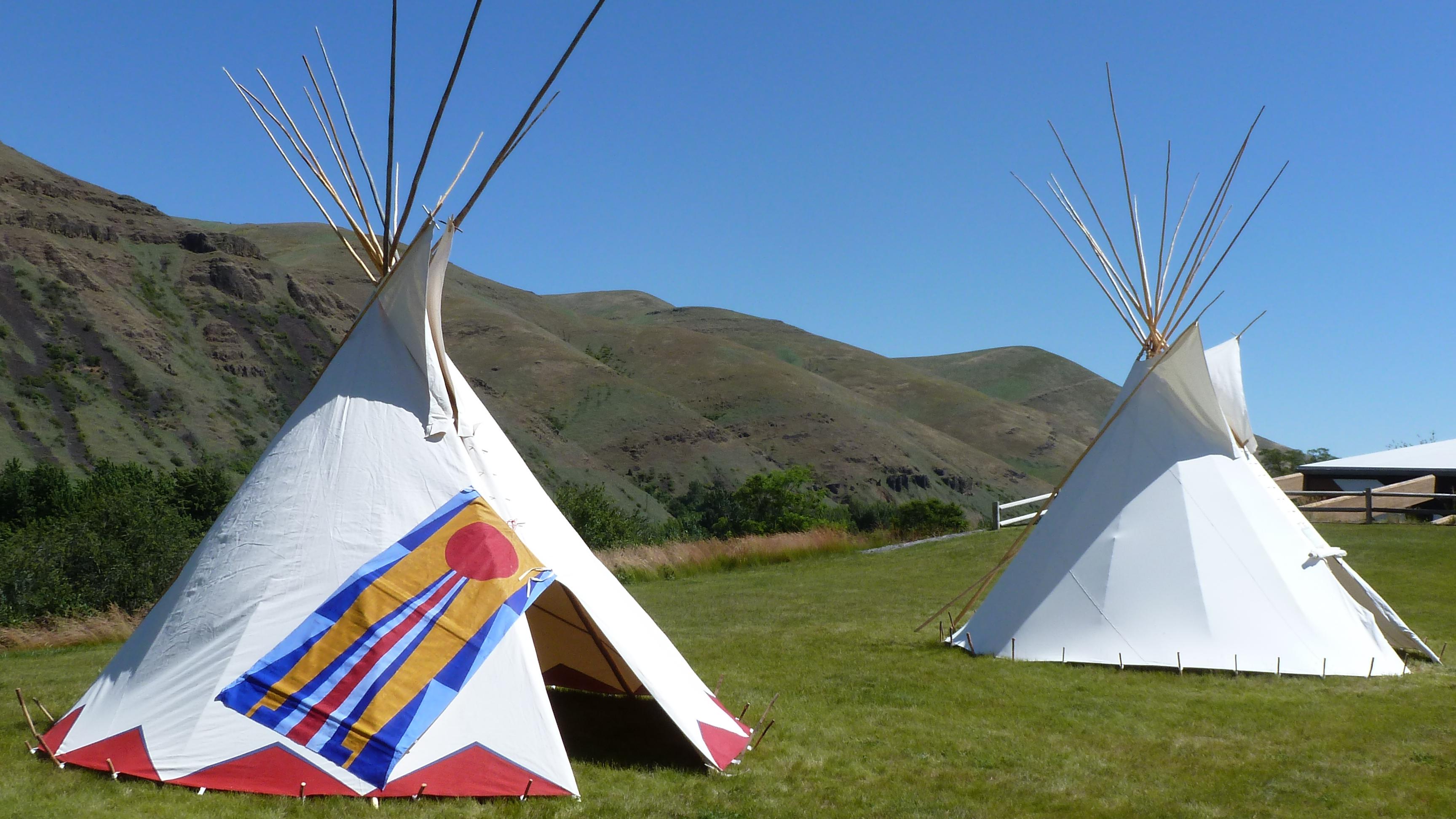 One painted and one plain tipi set up on fresh grass with hills and blue sky in background. 
