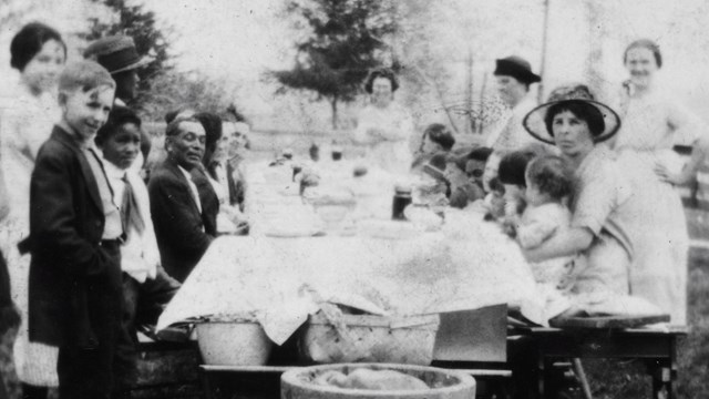 Black and white photograph of many people sitting on the sides of a long table full of food.