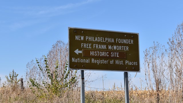 A brown historic site sign stands on a roadside. On it, a white arrow points left.
