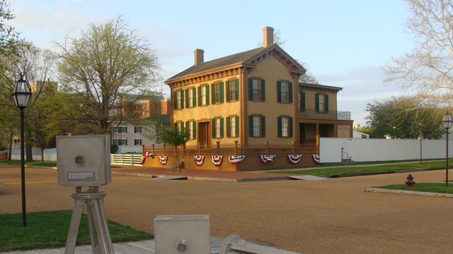 A low brick wall and wooden fence surround a two-story tan house with green window shutters.