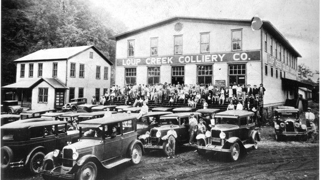 A black and white photo of a store with many people and old-fashioned cars in the foreground