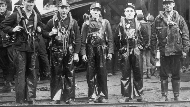 A black and white photo of miners wearing breathing equipment in front of a wooden structure