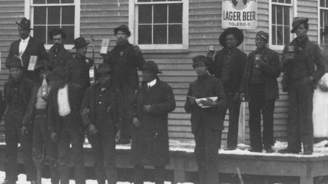 A black and white photo showing a group of African American men standing outside a saloon
