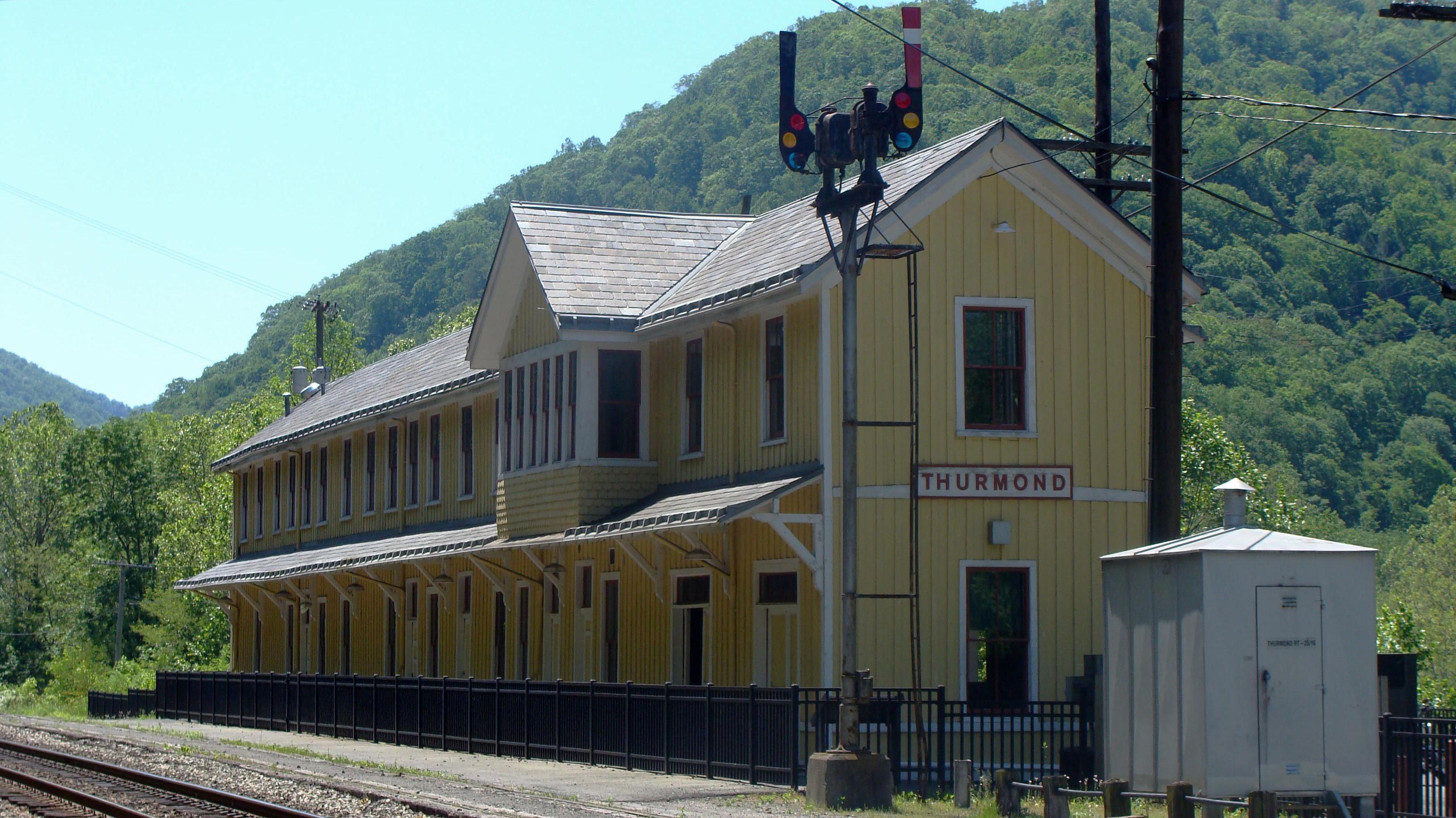 A long yellow wooden building with a sign that says Thurmond on it sits next to railroad tracks
