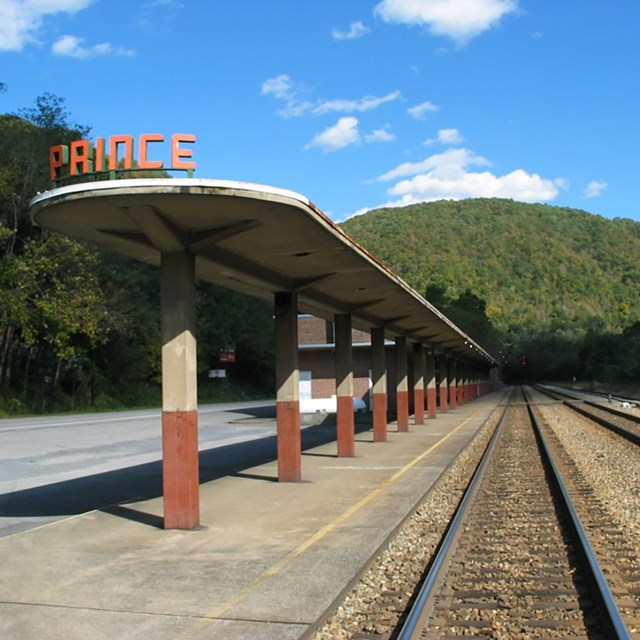 A train platform and depot next to railroad tracks. The depot has a sign that says Prince.
