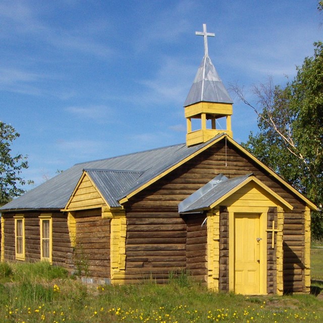 A log church with yellow trim. A grass and wildflower lawn.