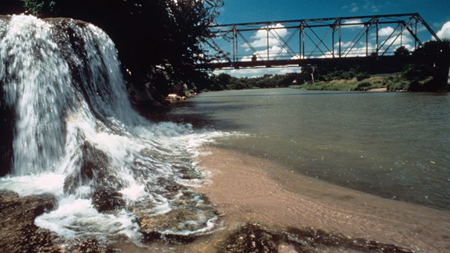 A waterfall flows into a river, a bridge behind it in the distance.