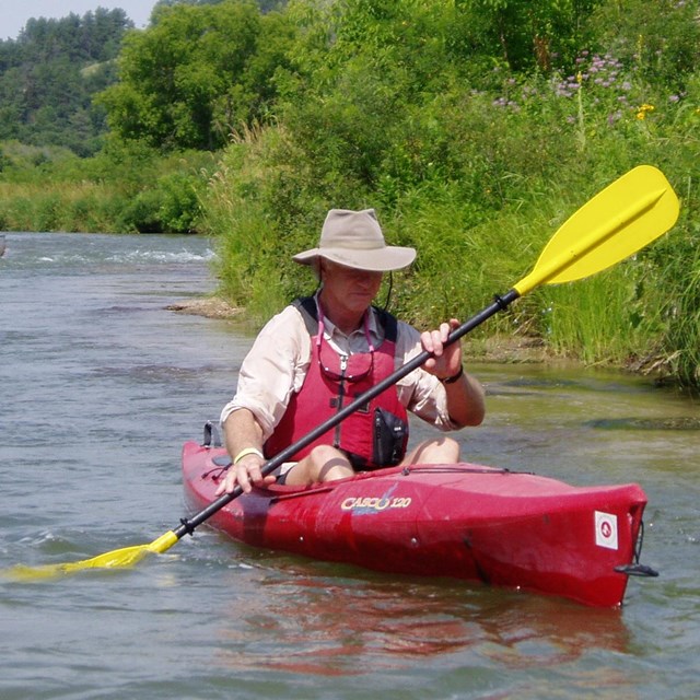 A kayaker in a red boat with a lifejacket and hat on