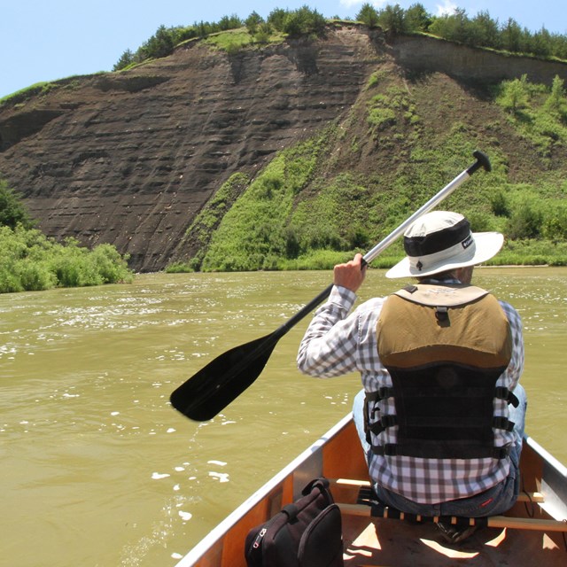 A canoer faces forward on a river, moving toward a dirt bluff