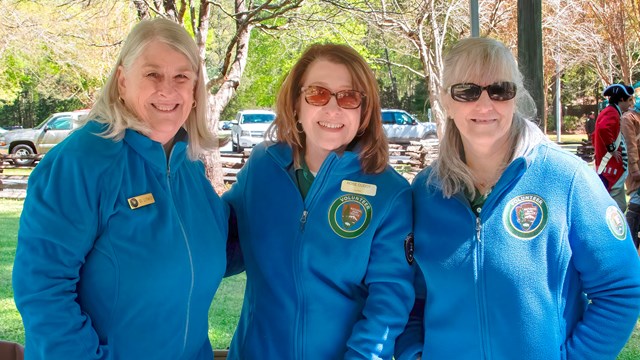 Three women in blue volunteer jackets stand at a children's table. 